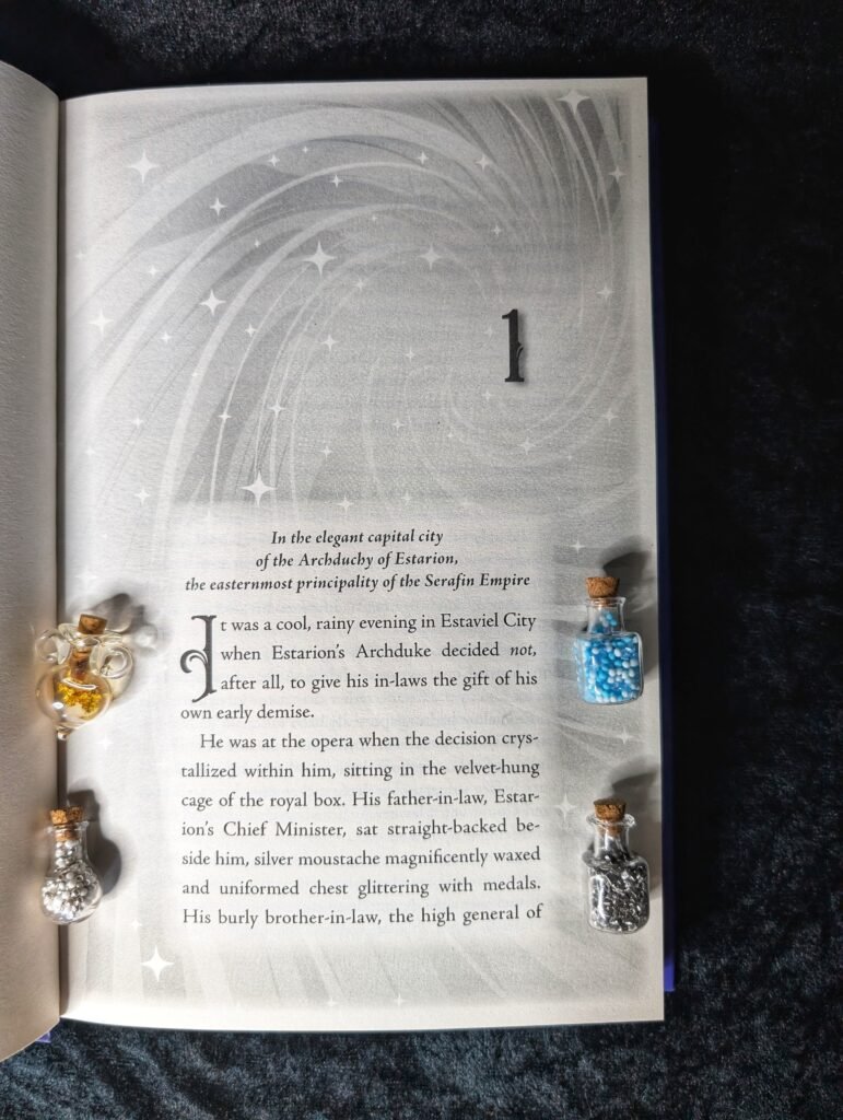An open book photographed from above, showing the start of chapter 1. 
The page has a soft grey background with swirling star-like patterns. Decorative glass bottles filled with glitter and beads are placed around the page.

Printed text reads:
“1
In the elegant capital city of the Archduchy of Estarion,
the easternmost principality of the Serafin Empire
It was a cool, rainy evening in Estaviel City when Estarion’s Archduke decided not, after all, to give his in-laws the gift of his own early demise.
He was at the opera when the decision crystallized within him, sitting in the velvet-hung cage of the royal box. His father-in-law, Estarion’s Chief Minister, sat straight-backed beside him, silver moustache magnificently waxed and uniformed chest glittering with medals. His burly brother-in-law, the high general of”
(The text cuts off at the bottom of the page.)